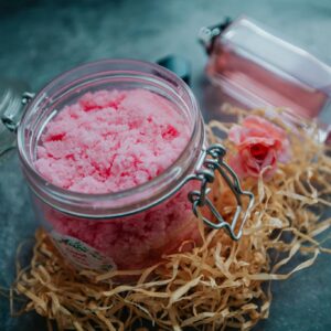 From above of pink body scrub in glass jar placed on table with perfume bottle and pile of shredded paper