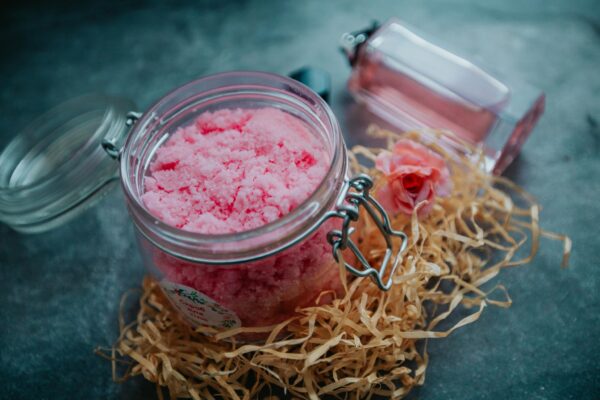 From above of pink body scrub in glass jar placed on table with perfume bottle and pile of shredded paper